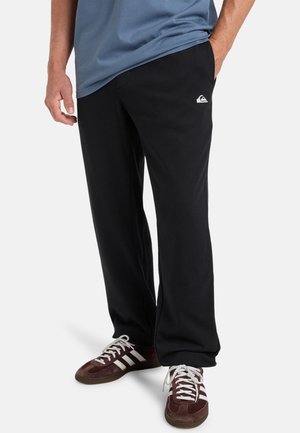 Pantalones de chándal negros en un ajuste relajado con un bolsillo lateral, combinados con zapatillas de deporte rojo oscuro con rayas blancas, llevados con una camiseta azul.