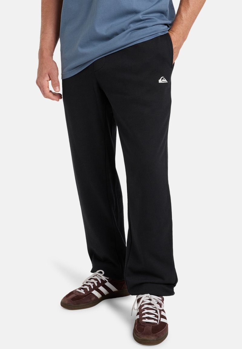 Pantalones de chándal negros en un ajuste relajado con un bolsillo lateral, combinados con zapatillas de deporte rojo oscuro con rayas blancas, llevados con una camiseta azul.