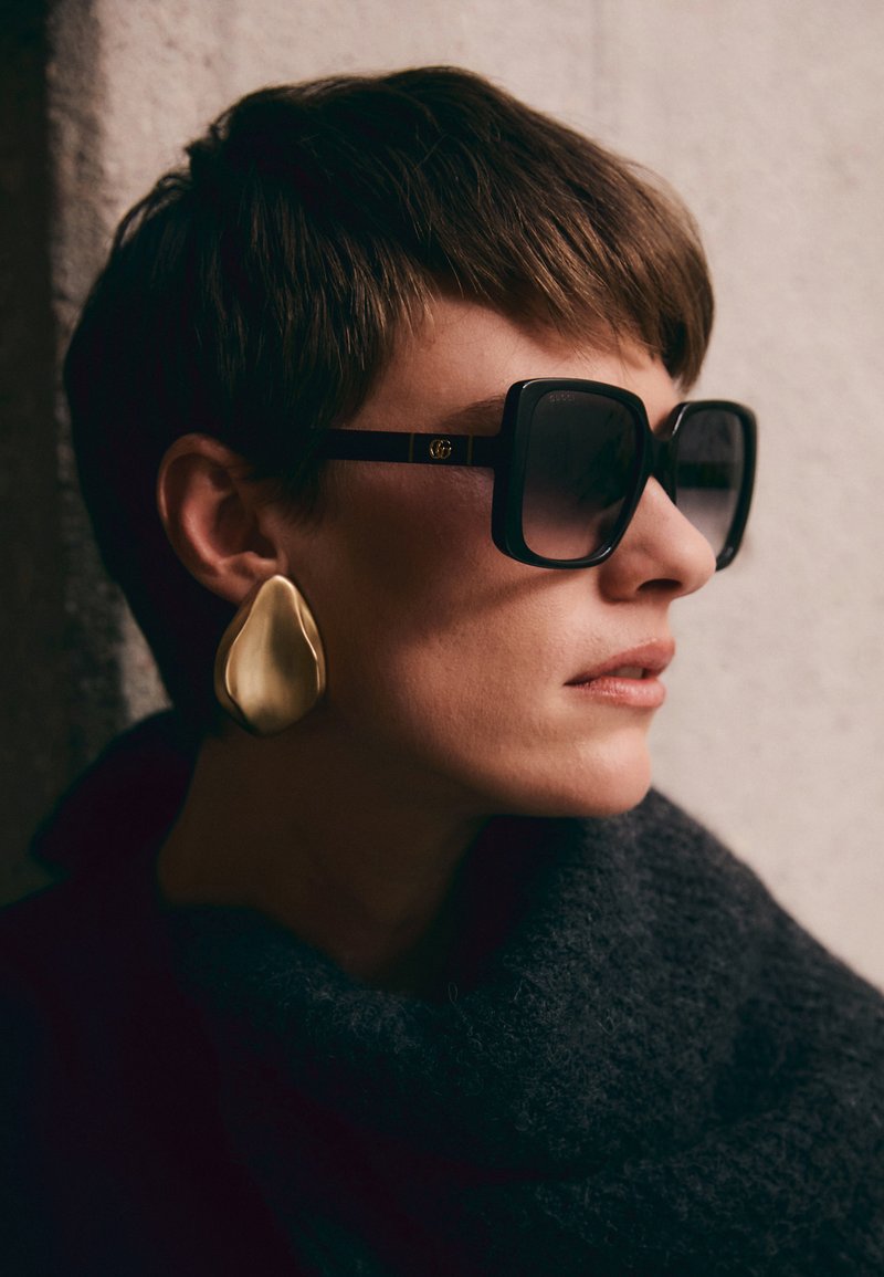 Gafas de sol negras de gran tamaño con lentes oscuros y detalles dorados, combinadas con un gran pendiente en forma de lágrima dorado y brillante. Bufanda texturizada oscura.