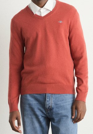 Homme portant un pull corail à col en V par-dessus une chemise blanche à col avec un jean bleu, debout devant un fond uni.
