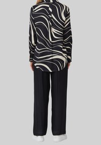 Camicia a maniche lunghe con un motivo ondulato in bianco e nero, abbinata a pantaloni neri larghi e sneakers bianche, su uno sfondo grigio.