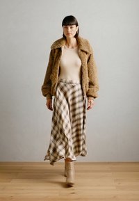 Fuzzy brun bomberjakke over en beige top, parret med en plaid maxi nederdel i beige og brune nuancer. Beige ankelstøvler.