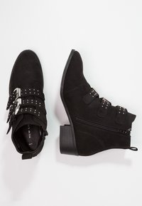 Botas de tobillo de ante negro con tres correas con tachuelas y hebillas plateadas, cremallera lateral y tacón cuadrado, mostradas desde vistas superior y lateral.