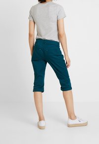 Turkusowe spódnice w wersji cropped z podwiniętymi mankietami, wykonane z elastycznego materiału; połączone z szarym t-shirtem z krótkim rękawem i białymi płóciennymi butami.