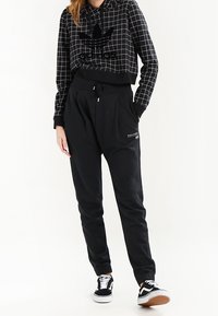 Mustat ruudulliset lyhyt cropped-hupparit, joissa on iso musta logo, yhdistettynä mustiin kapeneviin joggereihin. Mustat lenkkarit, joissa on valkoisia yksityiskohtia.