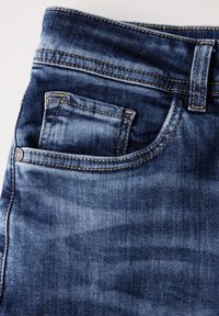 Jeans de mezclilla azul con una textura suave, que cuentan con un bolsillo frontal, presillas para el cinturón y costuras amarillas contrastantes. La tela tiene un efecto desgastado.