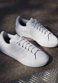 Par hvide Adidas sneakers med snørebånd og gult logo, placeret på en tekstureret betonoverflade i naturligt lys.