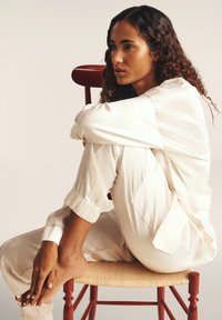 Jumpsuit blanc en satin avec des manches froncées et des poches, porté par une personne assise sur un tabouret tissé de couleur brun clair, devant un arrière-plan neutre.