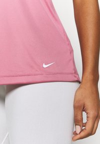 Růžový sportovní top Nike z hladké látky s jemným lemem a bílým logem Nike na levé straně, v kombinaci s bílými legínami.