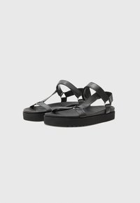 Sorte lær sandaler med flad sål, der har brede remme og en tåsløjfe. Glat tekstur, minimalistisk design og sølvhardware-detaljer.