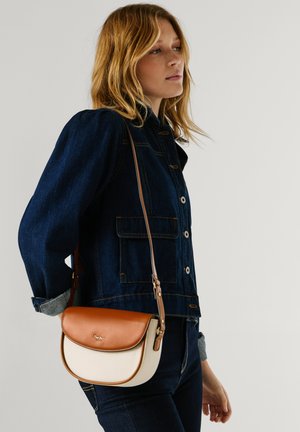 Žena u tamnoj traper jakni i trapericama, nosi smeđu i kremastu torbu na remenu s preklopnim zatvaračem.