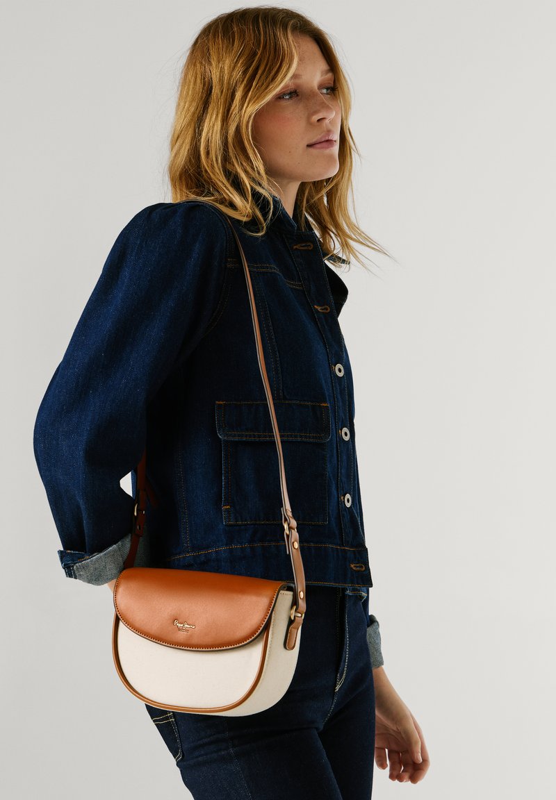 Vrouw met een donker denim jack en spijkerbroek, draagt een tweekleurige bruine en crèmekleurige schoudertas met klep sluiting.