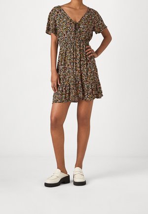 Femme portant une robe courte noire à motifs floraux avec un ourlet à volants et des mocassins blancs, debout avec une main sur la hanche devant un fond uni.