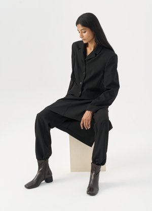 Kvinne med langt mørkt hår sittende, iført en løs svart blazer, matchende vide bukser og mørke ankelstøvler mot en ensfarget hvit bakgrunn.