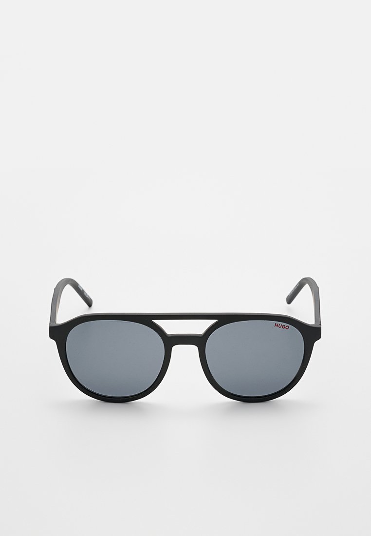 Schwarze Sonnenbrille mit grauen Gläsern, einem kantigen Design und einer Doppelbrücke. Sie hat dezente rote Markenbeschriftung am oberen linken Bügel.