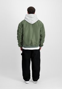Veste bomber verte avec des manches matelassées, une capuche grise et un ourlet côtelé. Portée par-dessus une chemise blanche et associée à un pantalon noir ample.