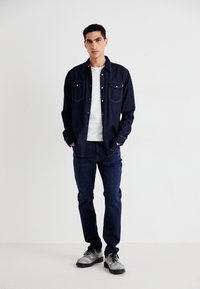 Chemise en denim marine avec deux poches poitrine, t-shirt blanc en dessous, associée à un jean sombre et des chaussures grises, présentant un look décontracté.