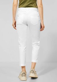 Kvinna som bär vita korta byxor med elastisk midja och beige sneakers, sedd bakifrån mot en grå bakgrund.