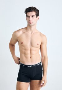 Fekete Nike boxer sort fehér logóval a derékpánton, testhezálló kialakítással és sima szövettextúrával. A modellen izmos testalkat van.