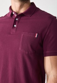 Bordový polo tričko s golierom na tri gombíky, s náprsním vreckom a malým pruhovaným akcentom v červenej, bielej a modrej farbe. Bavlnená textúra.