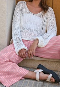 Pull blanc à maille ajourée avec manches longues ; pantalon ample rayé rose et blanc ; sandales plates noires à brides, assis sur une surface.