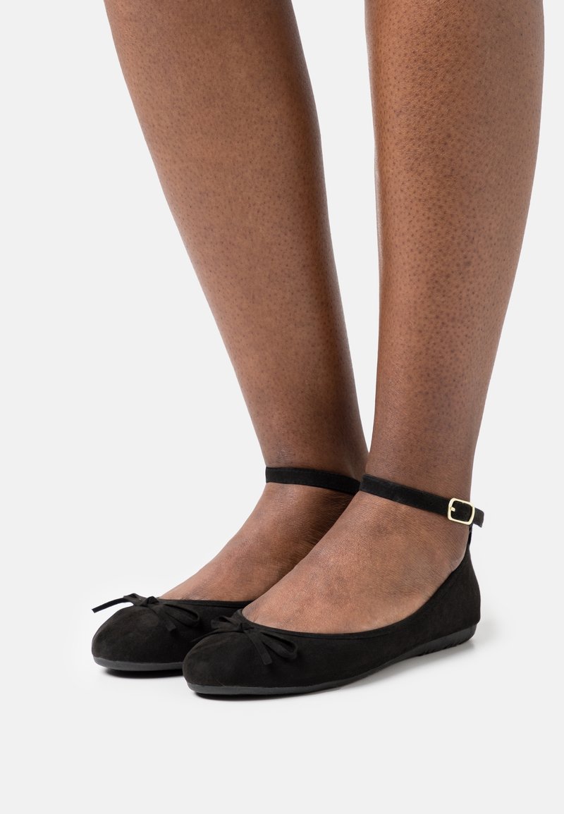 Ballerines en cuir nubuck noir avec un bout rond, un détail de nœud et une bride à la cheville dotée d'une boucle dorée. Texture lisse ; semelle plate basse.