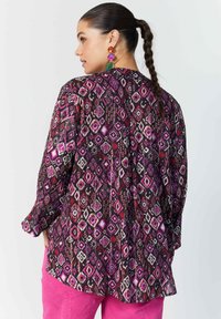 Bunt gemusterte Bluse mit einem Diamantdesign in Rosa-, Weiß- und Lilatönen auf einem schwarzen Hintergrund, mit langen Ärmeln.