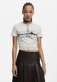 Svetlo béžové bavlnené tričko s čiernym potlačou púštnych krajín a textom. V kombinácii s tmavou plisovanou koženou sukňou. Ozdobný náhrdelník typu choker.