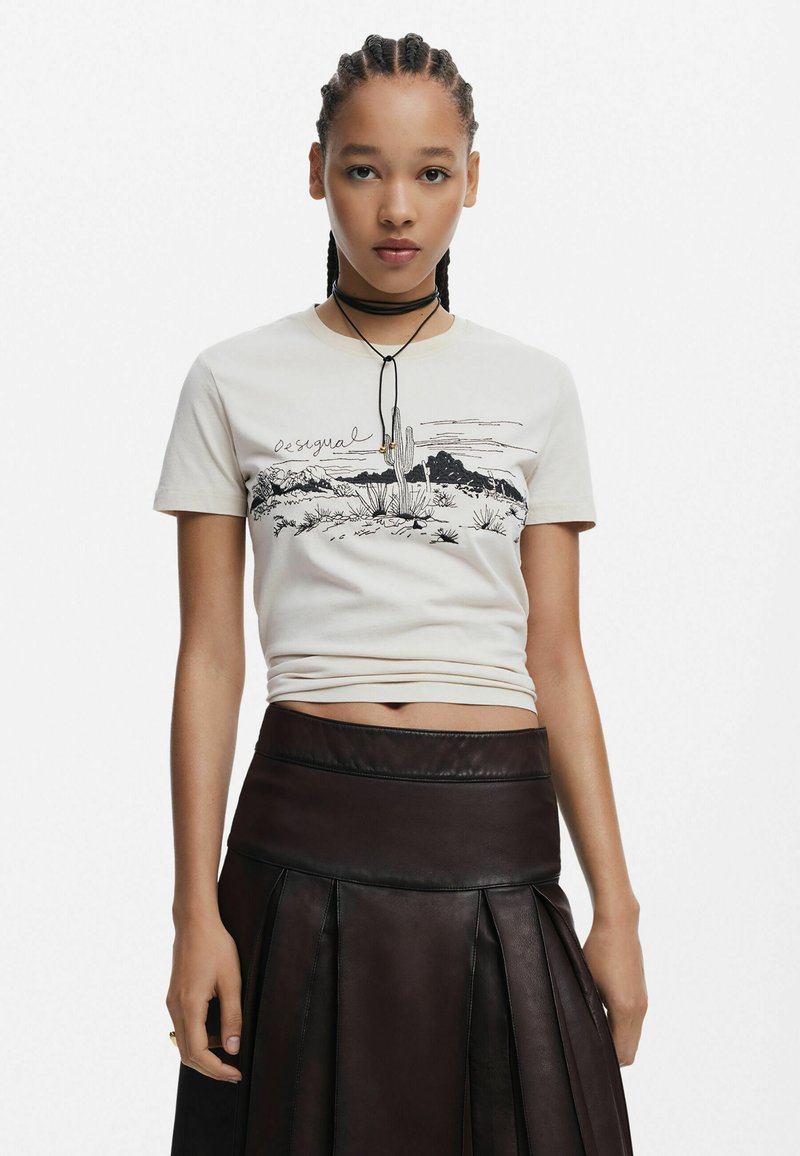 Svetlo béžové bavlnené tričko s čiernym potlačou púštnych krajín a textom. V kombinácii s tmavou plisovanou koženou sukňou. Ozdobný náhrdelník typu choker.