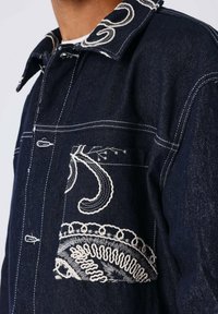 Donkerdenimjas met een gedetailleerd wit geborduurd ontwerp op de kraag en de borstzak, met contrasterende witte stiksels.