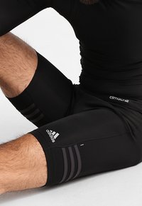 Pantalones cortos deportivos negros hechos de tejido elástico, con tres rayas gris oscuro a los lados y un logo de Adidas en el dobladillo.