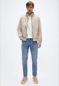 Μπεζ μπουφάν τύπου bomber με ψηλό γιακά, λευκό μπλουζάκι, ανοιχτόχρωμο τζιν και ταμπά αθλητικά παπούτσια. Μινιμαλιστικός σχεδιασμός με ομαλές υφές.