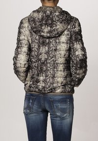 Vadderad jacka med ett ormskinsmönster i svart och krämfärg, med huva och elastiska ärmslut. Buren med blå denimjeans.