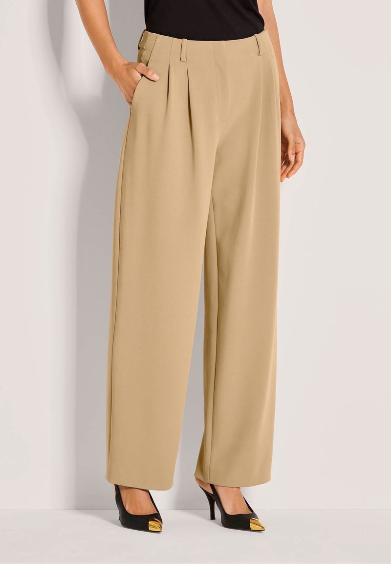 Beige wijde pantalon met voerplooien, gedragen met zwarte puntige hakken met gouden metalen accenten op de tenen.