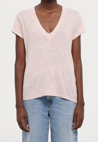 Jasnoróżowa koszulka z dekoltem w serek i krótkim rękawem, wykonana z miękkiego materiału. Charakteryzuje się luźnym krojem i lekko zaokrąglonym dołem. Nosi z niebieskimi dżinsami.