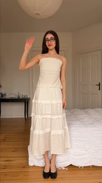 Jeune femme aux longs cheveux foncés et lunettes, portant une robe blanche bustier et des chaussures noires, debout et faisant signe dans une chambre.