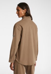 Ruskean kauluspaita, jossa on pitkät hihat, rento istuvuus ja hieman kaareva helma. Koristeluommel olkapäällä. Silkkinen kangas, jossa on miellyttävä rakenne.