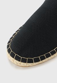 Mustat espadrillot, joissa on teksturoitu pinta, pyöreä kärki ja juuttiköysisohva. Reunassa olevat ompelemahat lisäävät kontrastista korostusta.