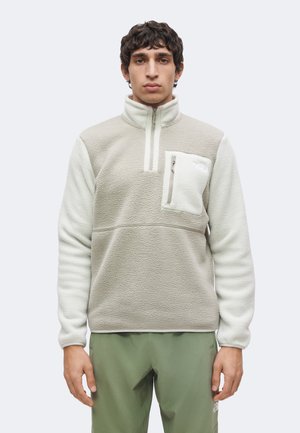Man staat recht vooruit, draagt een lichtbeige en witte fleece trui met een borstzak met rits en olijfgroene broek, neutrale achtergrond.