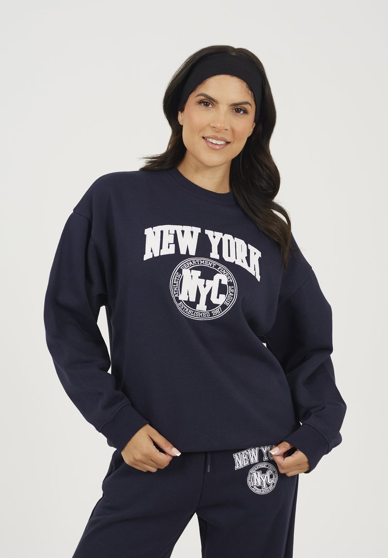 Donna che indossa una felpa blu marina e pantaloni con testo bianco "New York NYC", sorridendo mentre sistemava l'orlo della sua felpa.