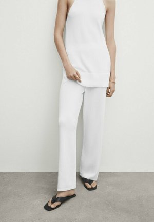 Woman wearing sleeveless white top and white wide-leg pants, standing on gray floor with black flip-flops.