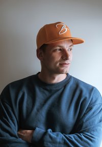 Narancssárga sapka, elöl fehér hímzett "B" betűvel, sötétkék pulóverhez párosítva, amely kerek nyakkivágással és hosszú ujjakkal rendelkezik.