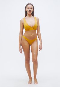 Σετ μπικίνι μουσταρδί με τριγωνικό μπραλέτο και ψηλόμεσο σλιπ, κατασκευασμένο από απαλό, ελαστικό ύφασμα.