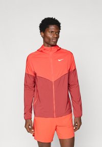 Άνδρας που φοράει ένα δίχρωμο κόκκινο ζακέτα Nike με κουκούλα και φωτεινές κόκκινες αθλητικές βερμούδες, που στέκεται μπροστά σε ένα απλό γκρίζο φόντο.