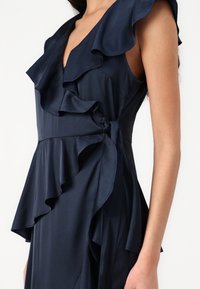 Vestido azul-marinho feito de tecido suave, com detalhes em babados ao longo do decote e do corpete, e um laço de amarração ao lado.