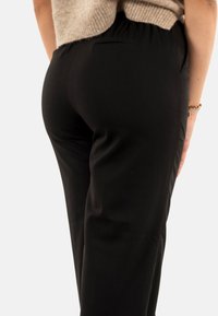 Pantalons noirs au toucher lisse, avec une taille élastique et des poches latérales. Le vêtement est ajusté et tombe droit jusqu'aux chevilles.