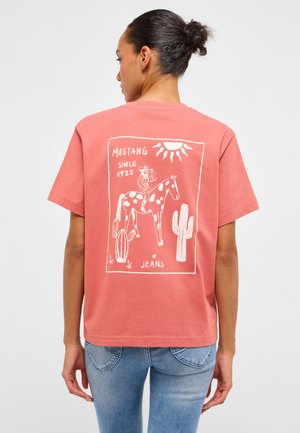 Woman with dark hair in a bun wearing a coral T-shirt with a white graphic of a spotted horse, cowboy, cactus, sun, and text "MUSTANG SINCE 1932 JEANS."