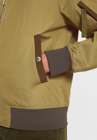 Main droite dans la poche d'une veste marron clair avec un revers, un bord de poche et une ceinture marron foncé, dotée d'un bouton-pression argenté sur la poche.