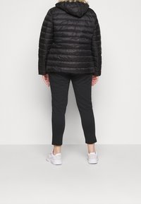 Giacca a piumino nera con cappuccio e bordo in pelliccia, indossata sopra pantaloni aderenti neri e sneakers bianche. Texture trapuntata con cuciture visibili.