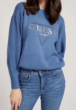 Donna che indossa un maglione di lana blu con il logo "GUESS EST. 1981" decorato con strass, abbinato a jeans blu a vita alta.